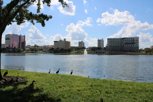 A curved colonnade and promenade along the edge of a city lake, palms and low buildings beyond.