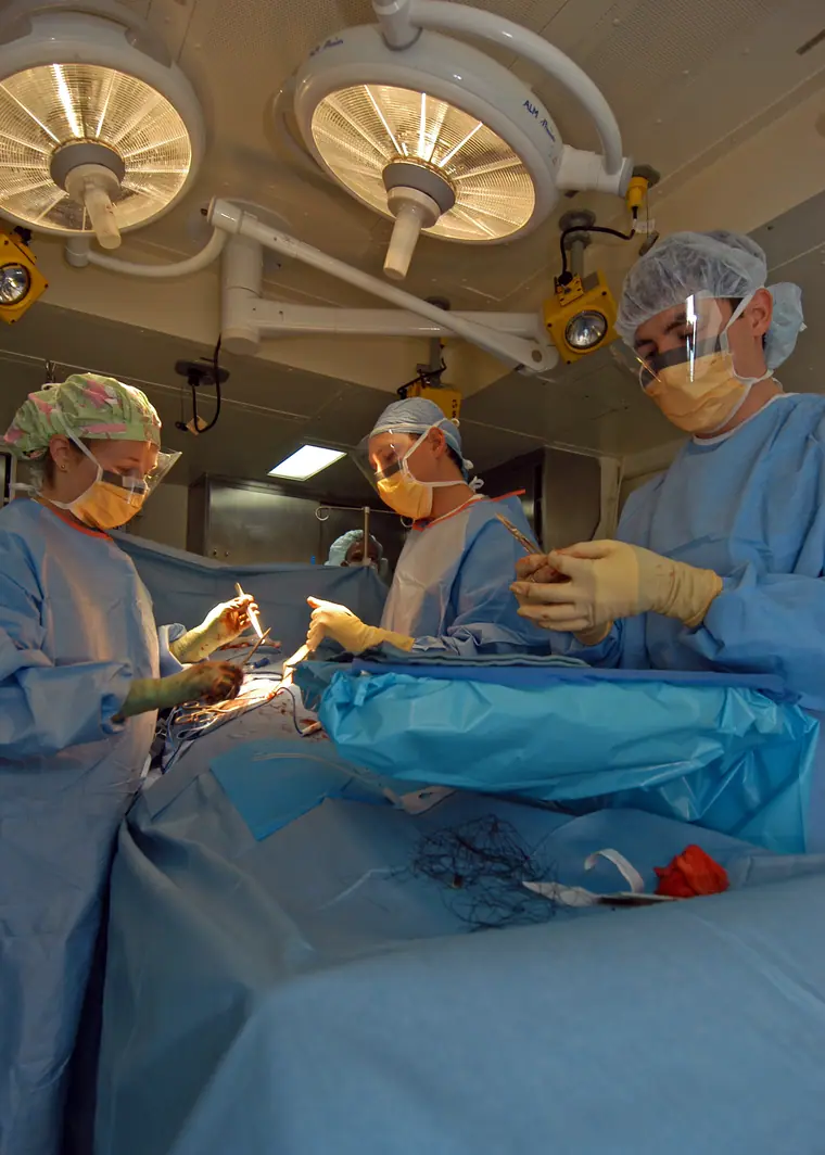 Three gowned and masked medical staff working over a draped operating table under surgical lights.
