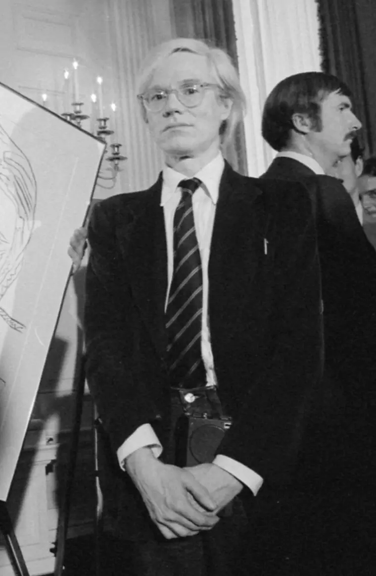 Andy Warhol in a dark jacket at a formal reception, white hair swept back, looking slightly off camera.