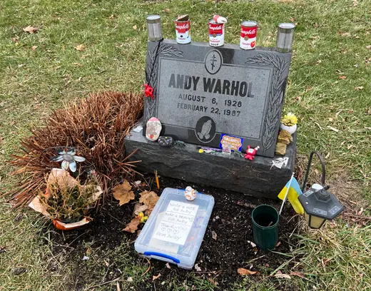 A modest granite headstone reading Warhola in a small hillside cemetery, with offerings left at its base.