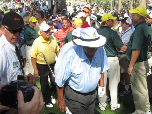 Three elderly golfers on a course during a par-three tournament, crowds behind them.