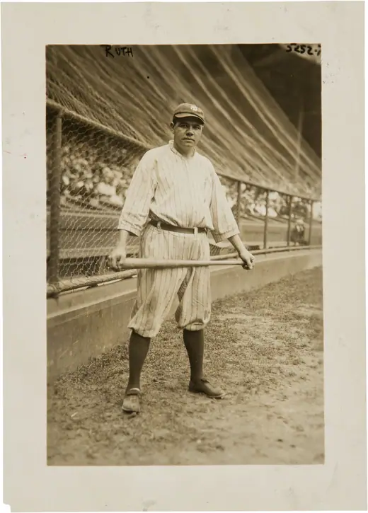 Ballplayer in a dark cap and uniform standing with a bat over one shoulder.