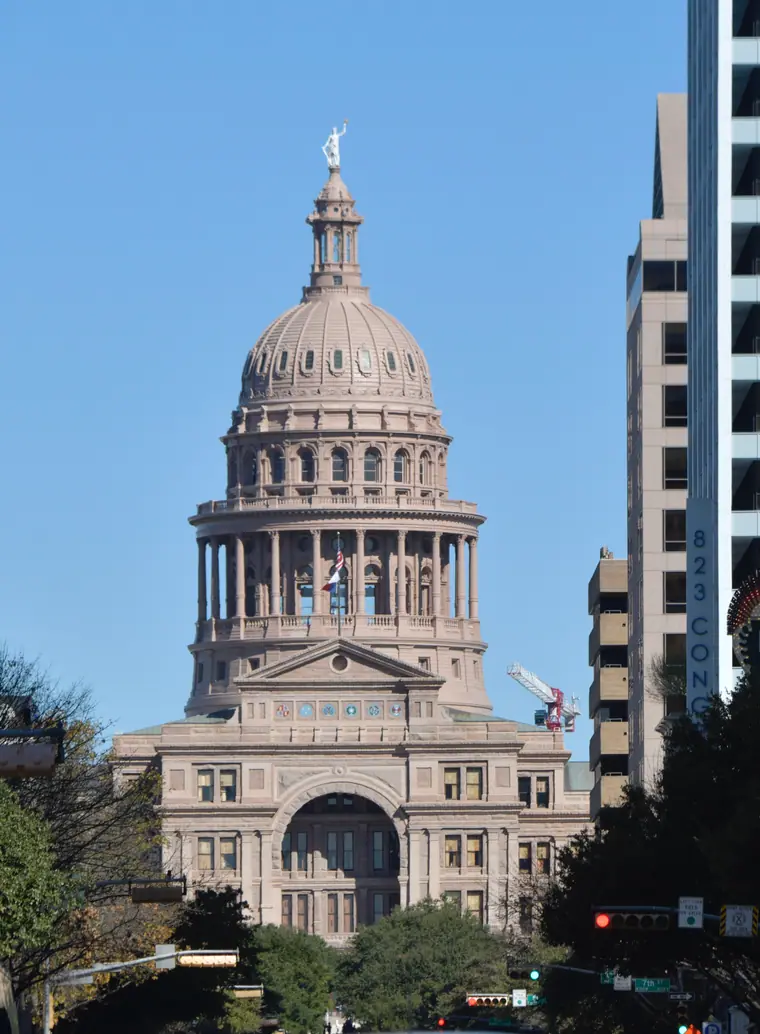The pink granite Texas State Capitol in Austin rising behind trees, its dome against a bright sky.
