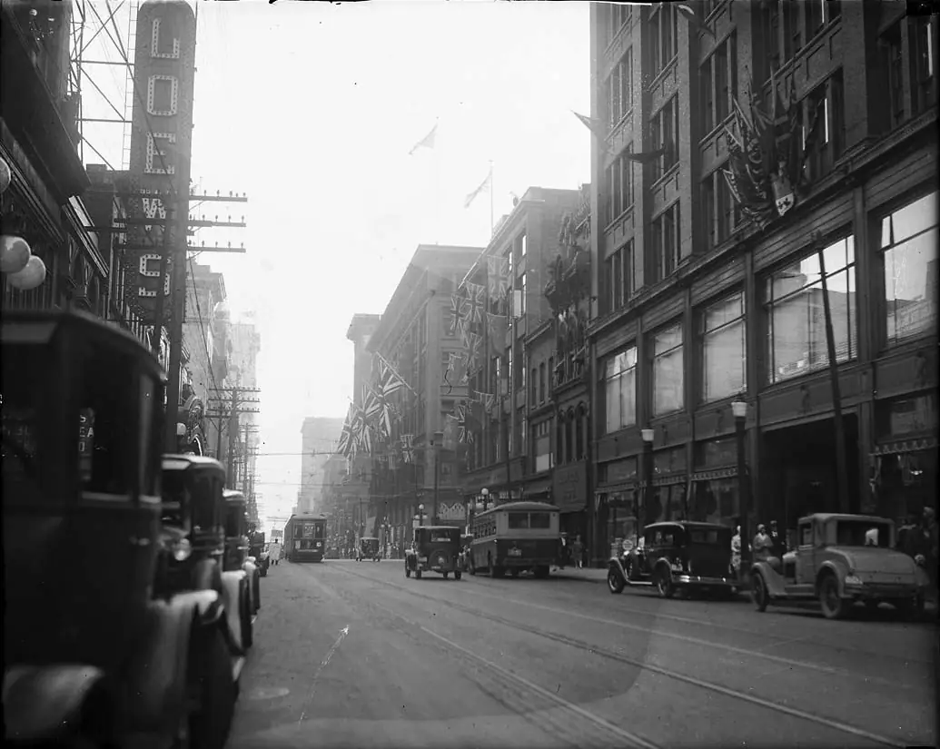 Busy Depression-era commercial street with streetcars, period cars, awnings and crowded sidewalks.