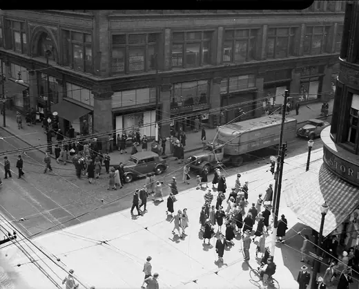 Street corner outside a large department store, streetcars turning and shoppers crossing.