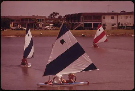 A small sailboat on a flat lake beside low apartment buildings in a Florida retirement community, photographed in 1973.