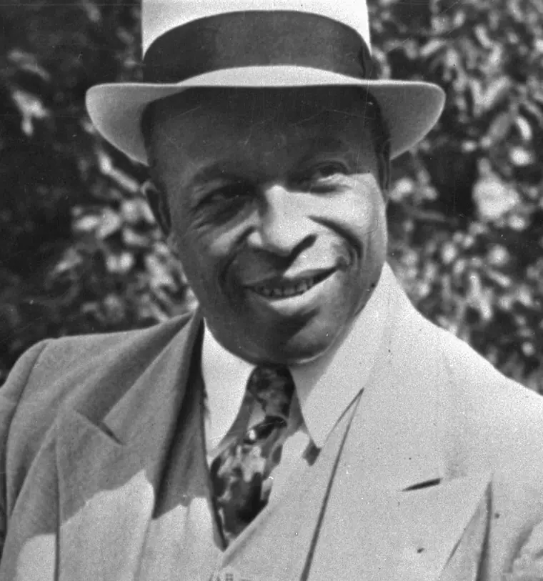 Press photograph of Father Divine in a suit, photographed in 1938 beside a sign at the Crum Elbow estate in Dutchess County, New York.