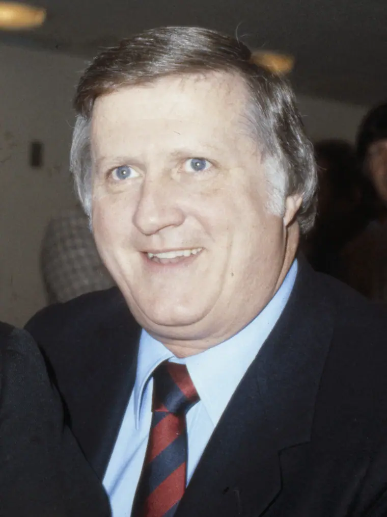 Black-and-white photograph of a heavy-set man in a dark blazer and open-collared shirt, caught mid-sentence.