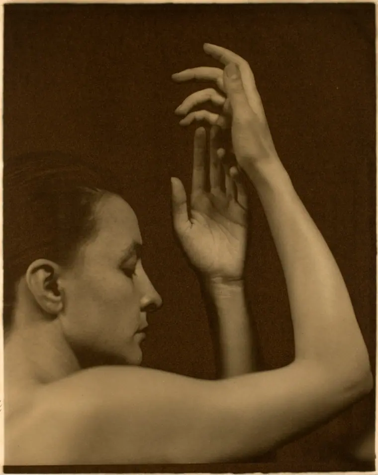 Black-and-white platinum print portrait of Georgia O'Keeffe, shoulders and hands raised, gaze lowered.