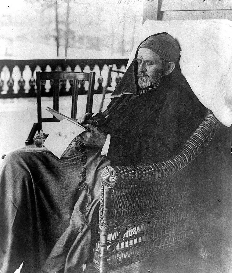 Ulysses S. Grant seated in a rattan chair on a porch, wrapped in a shawl with a cap on, writing on a board across his lap.