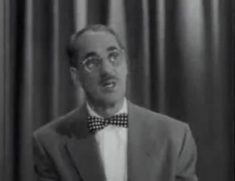 A television quiz-show host in a suit, seated behind a desk with a microphone, circa 1952.
