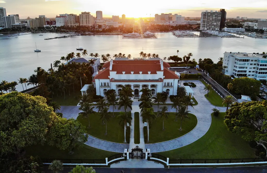 A white marble mansion with a colonnaded portico behind a lawn and palms.
