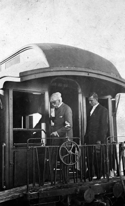 An elderly man in a light suit and hat standing on the open rear platform of a railway carriage.