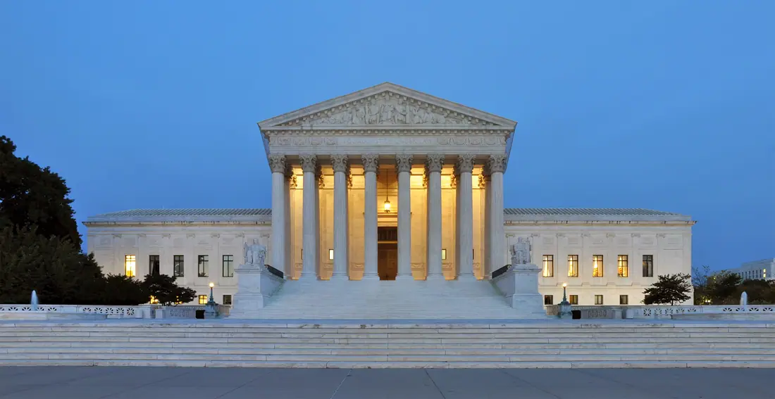 The west facade of the United States Supreme Court Building lit at dusk, with its columned portico and broad front steps.