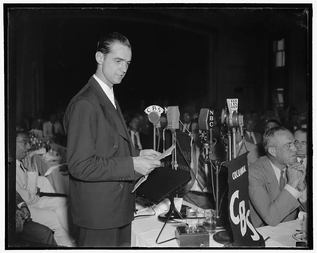 Man at a bank of radio microphones addressing a seated audience of men in suits at a press event.