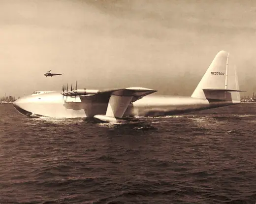Enormous eight-engined wooden flying boat sitting on water, dwarfing the small craft alongside it.