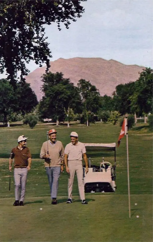Colour postcard of a low desert resort hotel behind a green golf course with palm trees.
