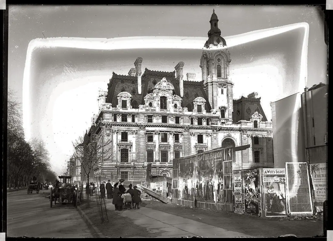 Enormous Beaux-Arts stone mansion with towers and a mansard roof on a snowy city corner, horse traffic in the street.