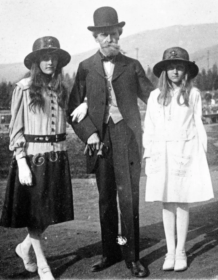 Elderly bearded man in a dark suit standing outdoors with two young girls in white summer dresses.