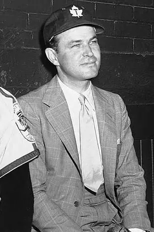 Man in a suit and tie seated in a baseball dugout in the mid-1950s.