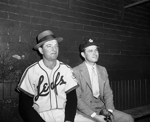 Two men talking in a baseball dugout, one in a suit, one in uniform.