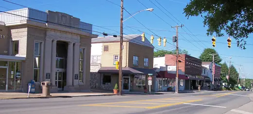 A short main street of low brick storefronts in a small town, hills rising behind.