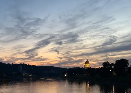 Twilight view along a wide river with a city skyline and an illuminated gold capitol dome at right.