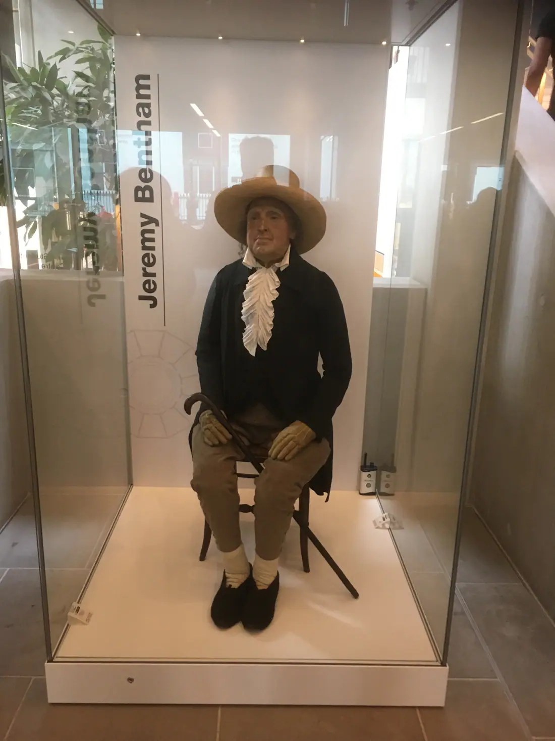 A seated figure in an early nineteenth-century black suit and wide-brimmed hat, holding a walking stick, displayed inside a glass case.