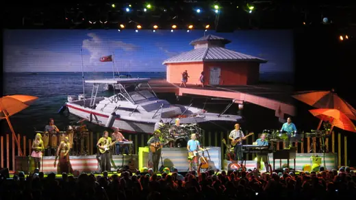 Band on a brightly lit arena stage with a large video screen behind them and a full crowd in front.