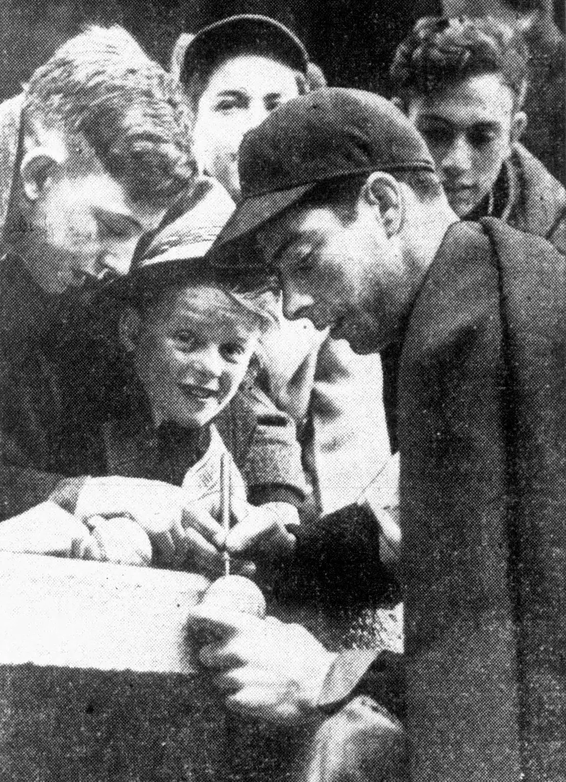 A ballplayer in uniform bent over a page, signing his name while a crowd of people watch.