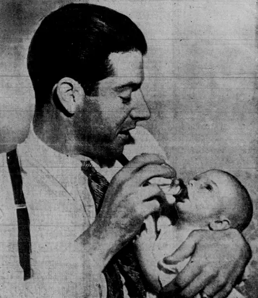 A man in a baseball uniform holding an infant, both looking toward the camera.