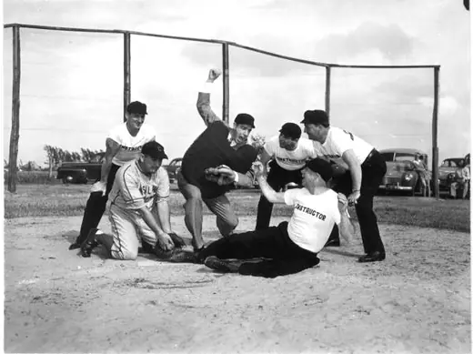 A ballplayer in a light suit standing with four men in dark umpire uniforms on a grass field.