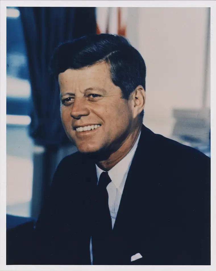 Colour portrait photograph of President John F. Kennedy in a dark suit, seated and looking toward the camera.