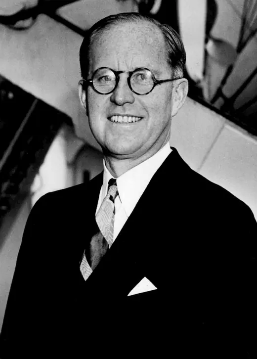 Photographic portrait of Joseph P. Kennedy Sr. in a light suit and round spectacles, smiling slightly.
