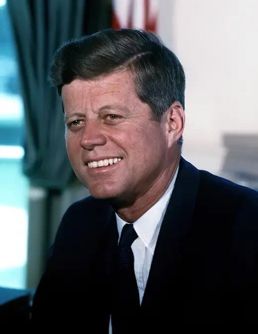 Colour photograph of John F. Kennedy in the Oval Office, standing in a dark suit.