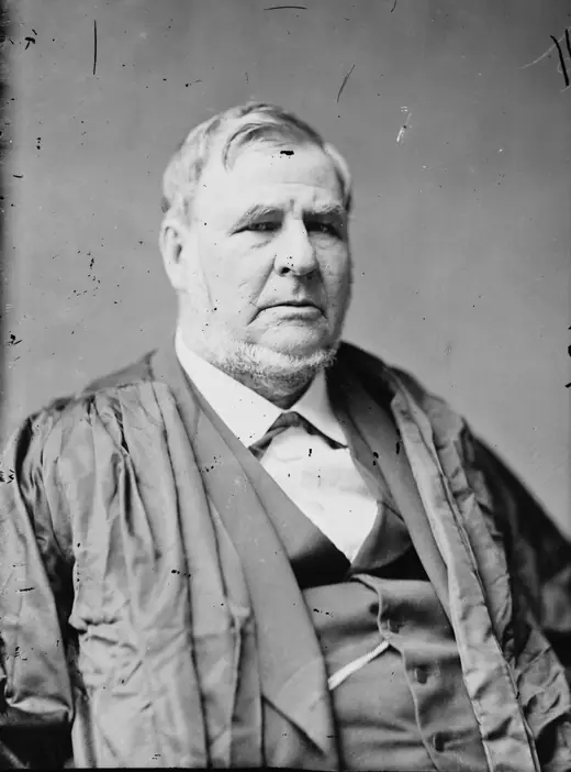 Studio photograph of a heavy-set bearded man in a dark suit, seated with one hand resting on a table.