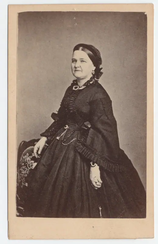 Carte de visite portrait of Mary Todd Lincoln standing in a wide hooped gown with a floral headdress.