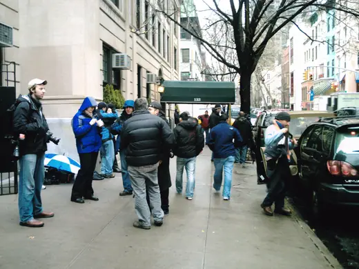 Press photographers with long lenses waiting on a Manhattan sidewalk outside an apartment building.