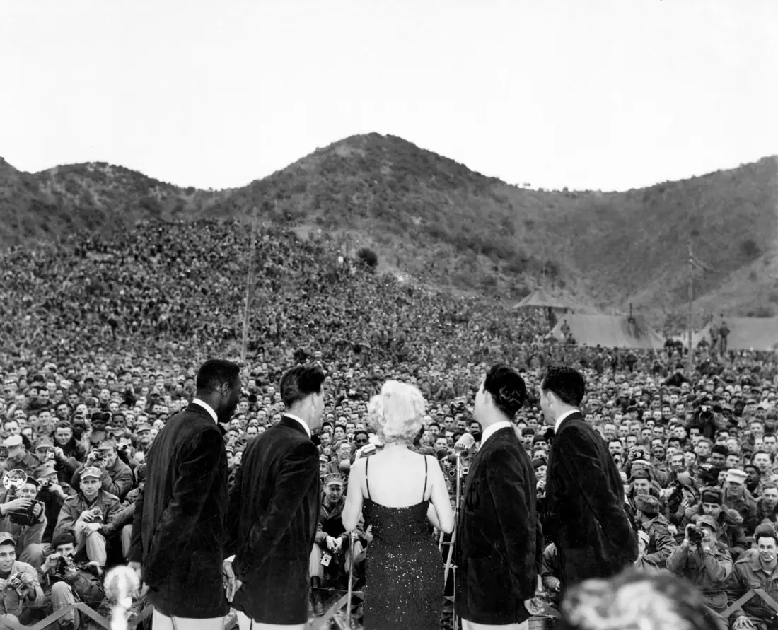 Woman in a dark dress singing on an outdoor stage before a hillside packed with thousands of uniformed troops.