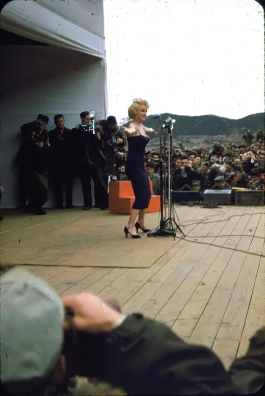 Marilyn Monroe on a bare wooden stage in a sleeveless dress, microphone in hand, soldiers visible below her.