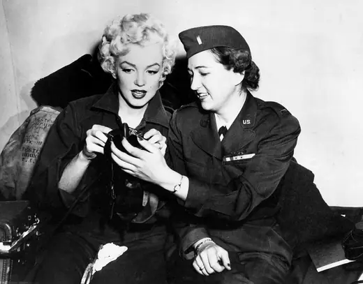Monroe in a flight jacket seated in a military aircraft cabin, smiling toward the camera.