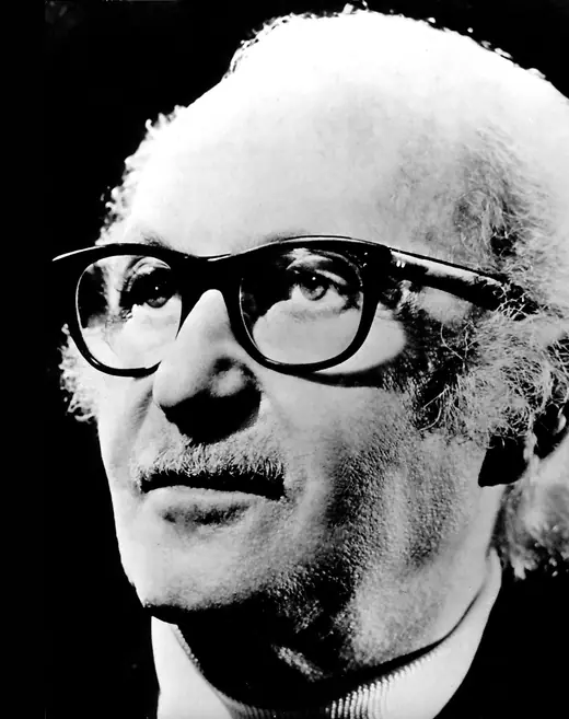 Head-and-shoulders photograph of an elderly bespectacled man in a suit and open-collared shirt.