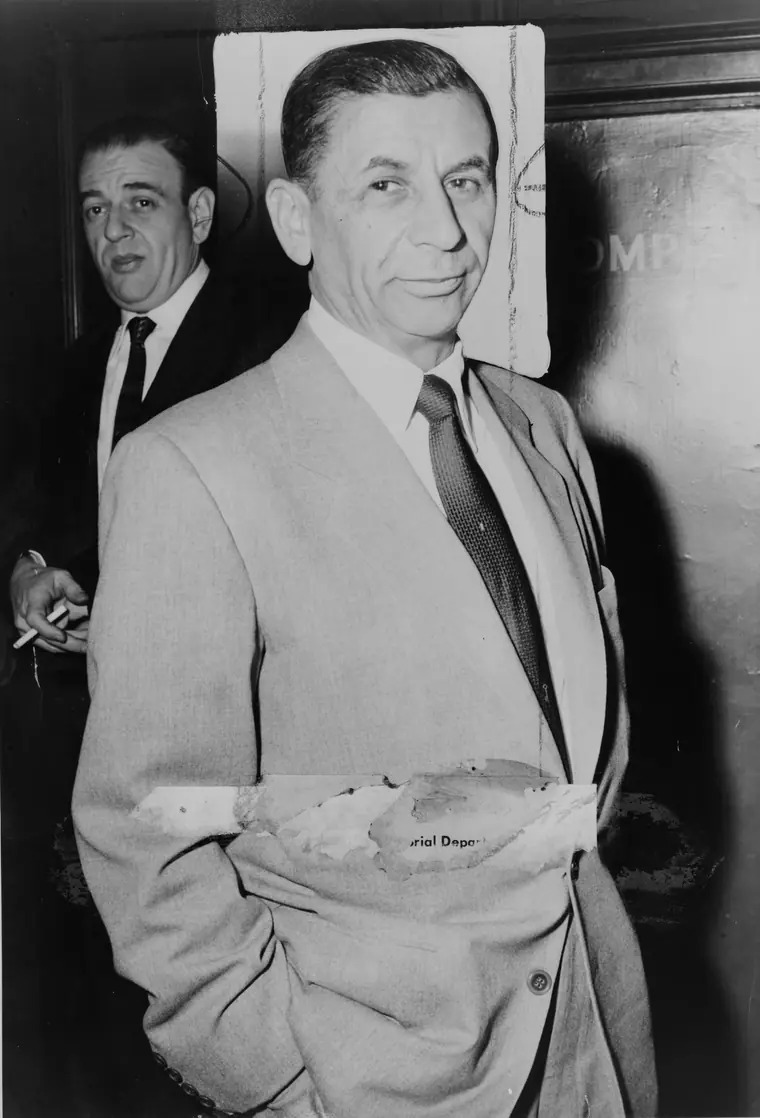 Black-and-white photograph of a small, older man in a dark overcoat and hat standing in a police station, looking toward the camera.