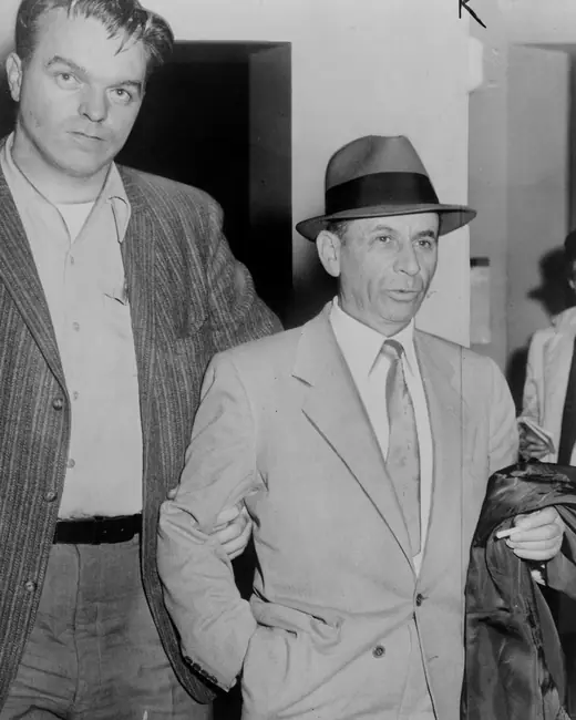 A man in a hat and overcoat being escorted by a detective into a New York police station.