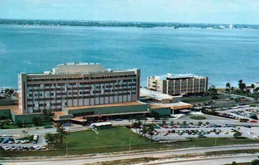 Colour postcard of a low white hospital complex on Miami Beach with palm trees and parked cars.