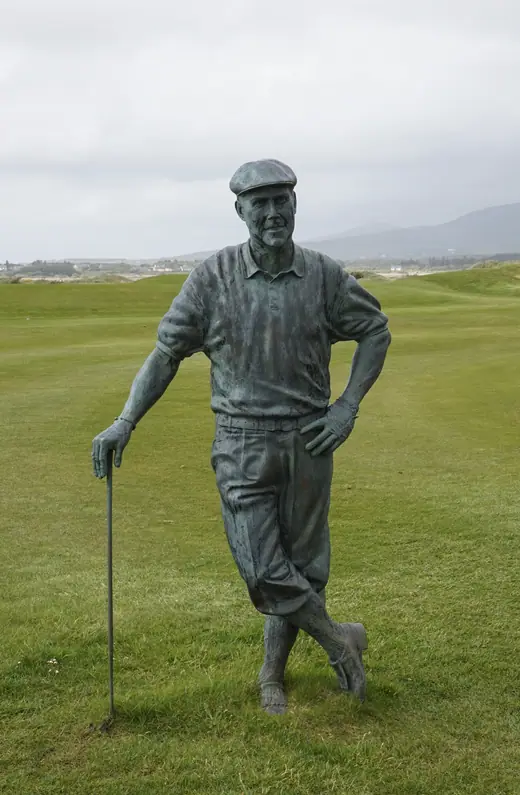 A bronze statue of a golfer in a flat cap completing a swing, on a links course by the sea.