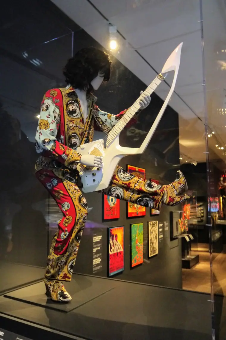 Museum display of a cherub-patterned stage suit on a mannequin holding a sculpted white guitar.
