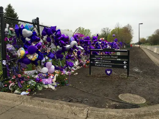 Gated entrance to a studio compound with purple ribbons and tributes tied along the fence.