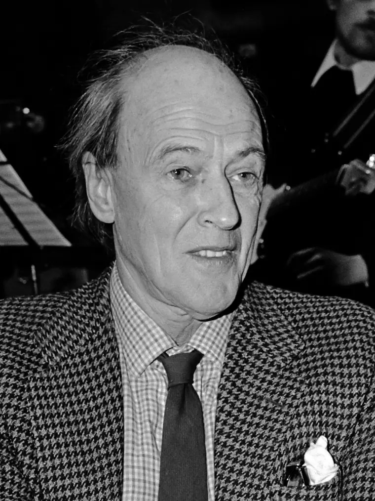 Roald Dahl in late middle age, seated at a press event, hands clasped, listening.