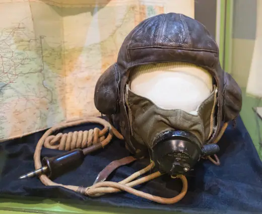A worn brown leather RAF flying helmet with oxygen mask and communication cables, displayed in a museum case.